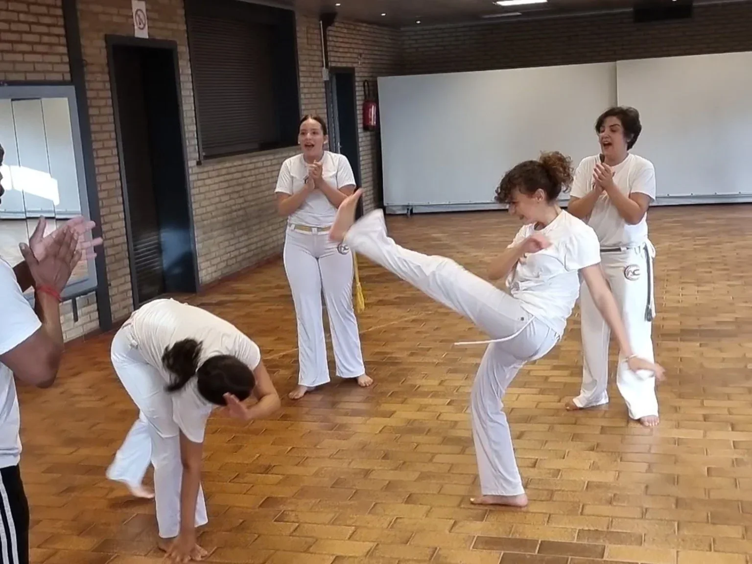 Twee capoeiristas spelen met een hoge trap en een ontwijking, terwijl anderen in de groep klappen.